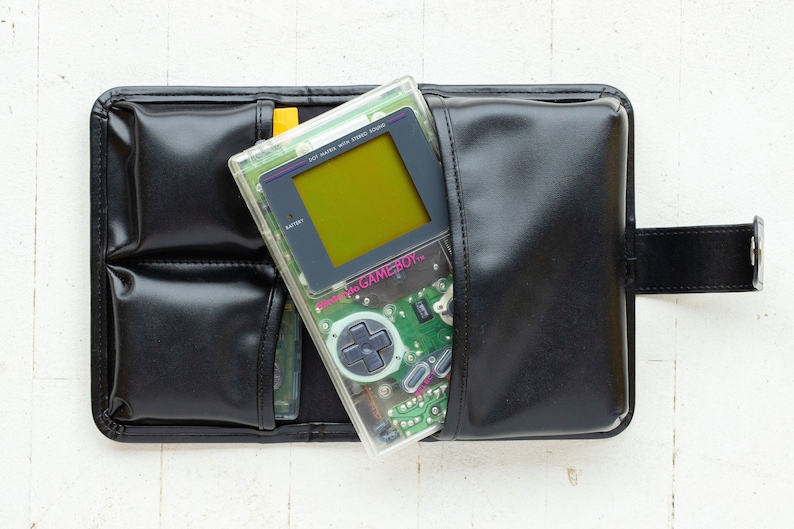 May include: A clear Game Boy Color handheld video game console with a green circuit board and a yellow screen. The console is in a black leather carrying case with multiple pockets.
