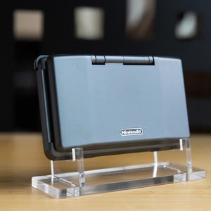 May include: A grey Nintendo DS Lite handheld console, displayed on a clear acrylic stand. The console is closed, with the Nintendo logo visible. The background is blurred, with a wooden surface in the foreground.