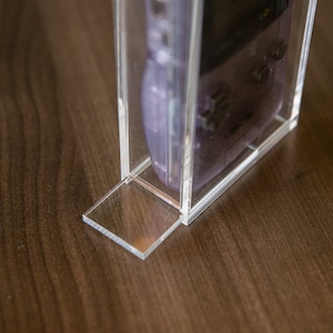 May include: A clear acrylic display case with a purple handheld video game console inside. The case has a clear base and a clear lid.