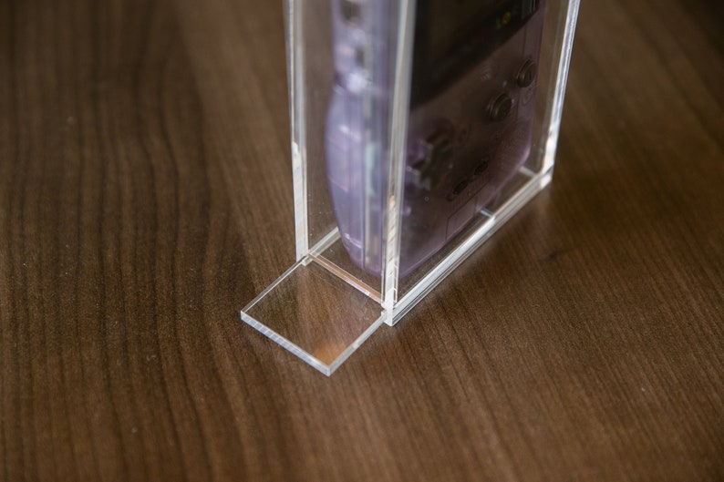 May include: A clear acrylic display case with a purple handheld video game console inside. The case has a clear base and a clear lid.