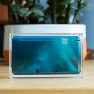 May include: A teal handheld gaming console enclosed within a clear acrylic display case. The console features a glossy finish with two small circular camera lenses visible on the outer shell. The transparent case provides a protective housing.