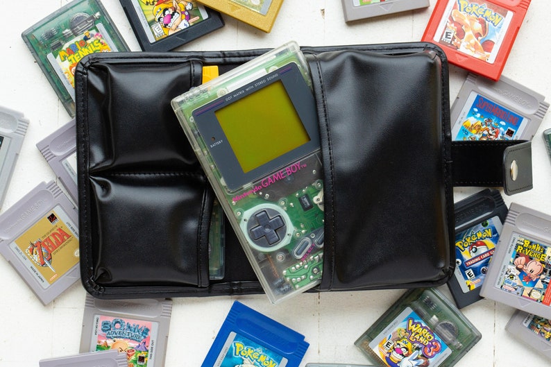 May include: A clear Game Boy Color handheld gaming console with a green screen and a black and white button pad. The console is in a black leather case with multiple slots for game cartridges. The case is surrounded by various Game Boy Color cartridges, including Pokemon, Zelda, and Sonic.