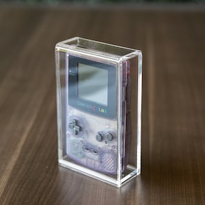 May include: A clear acrylic display case with a purple Game Boy Color inside. The Game Boy Color has a black screen and a gray and white design.