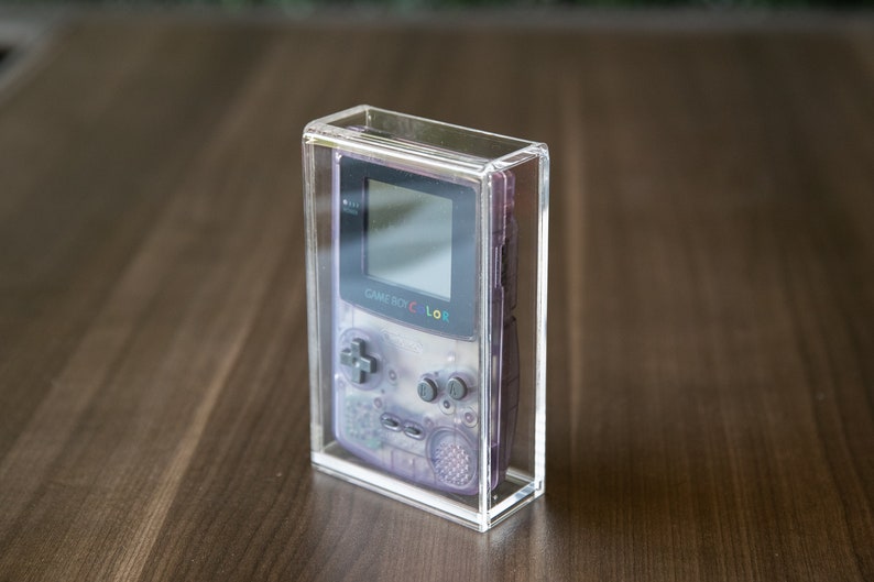 May include: A clear acrylic display case with a purple Game Boy Color inside. The Game Boy Color has a black screen and a gray and white design.