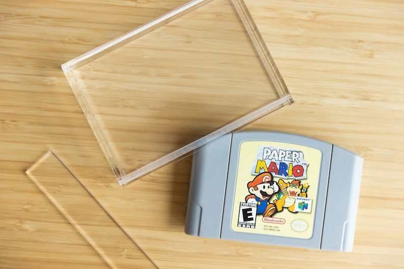 May include: A gray Nintendo 64 video game cartridge labeled "Paper Mario" with a colorful illustration of Mario and other characters. A clear acrylic display case is next to the game on a wooden surface.
