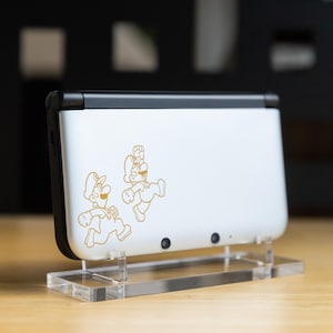 May include: A white handheld gaming console with a black top and sides, featuring gold-colored cartoon characters. The console is displayed on a clear acrylic stand, set against a blurred background.