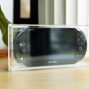 May include: A black Sony PS Vita handheld gaming console in a clear acrylic case. The console has a screen, buttons, and joysticks. The text "PS Vita" is visible on the console.
