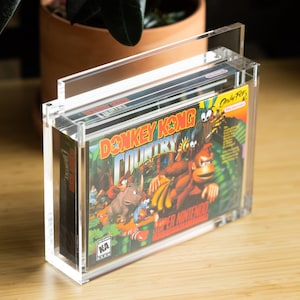 May include: A clear acrylic display case with a handle, showcasing a vintage Donkey Kong Country video game cartridge for the Super Nintendo Entertainment System. The game cartridge is in its original box with a colourful design featuring Donkey Kong and other characters.