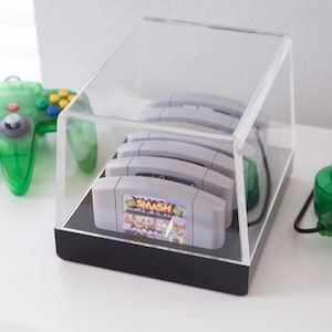 May include: A clear acrylic display case with a black base, holding a stack of five grey Nintendo 64 game cartridges. The top cartridge has a colourful label with the text "Super Smash Bros."