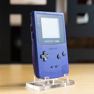 以下が含まれることがあります： 透明なアクリルスタンドに置かれた紫色のゲームボーイカラー携帯ゲーム機。画面は空白で、コンソールには方向パッド、AボタンとBボタン、そして「GAME BOY COLOR」と「Nintendo」の文字があります。