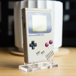 以下が含まれることがあります： 淡いグレー色のビンテージ任天堂ゲームボーイ携帯ゲーム機。画面、方向パッド、ボタン、そして「Nintendo GAME BOY」の文字があります。透明なアクリルスタンドに展示されています。