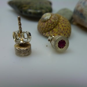 Könnte beinhalten: Nahaufnahme von silbernen Ohrsteckern mit einem kleinen, runden, roten Edelstein. Die Ohrringe liegen auf einer weißen Oberfläche, mit einer Muschel und Kieselsteinen im Hintergrund. Schlichtes, elegantes Design.