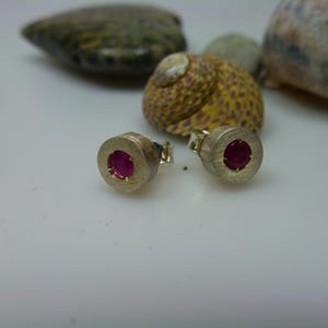 Könnte beinhalten: Ein Paar silberner Ohrstecker mit gebürsteter Oberfläche. Jeder Ohrring hat einen runden, leuchtend roten Edelstein in der Mitte. Die Ohrringe werden auf einer weißen Oberfläche mit Muscheln im Hintergrund präsentiert.
