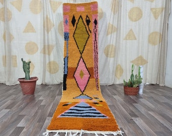 May include: A long, narrow rug with a geometric pattern in shades of pink, yellow, blue, and brown. The rug has a fringe border and is laid out on a wooden floor.