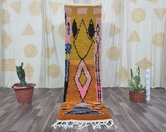May include: A long, narrow rug with a geometric pattern in shades of pink, yellow, blue, and black. The rug has a fringe on the bottom.