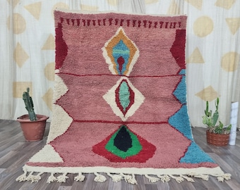 May include: A handmade, pink Moroccan rug with geometric designs. The rug features diamond and lozenge shapes in white, red, green, blue, and yellow. It has a fringe along the bottom edge and is displayed against a neutral backdrop.
