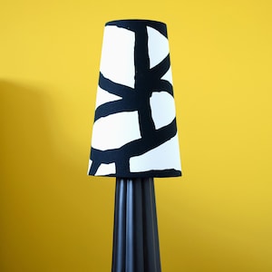 May include: A black table lamp with a white lampshade featuring a bold black geometric pattern. The lamp has a fluted base and sits on a wooden table. A book is visible on the table. The background is a solid yellow.