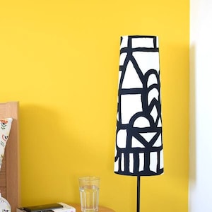 May include: A tall floor lamp with a cylindrical shade featuring a bold black and white geometric pattern. The lamp stands on a thin black metal pole next to a small wooden side table holding a clear glass of water and a book titled Lucky.