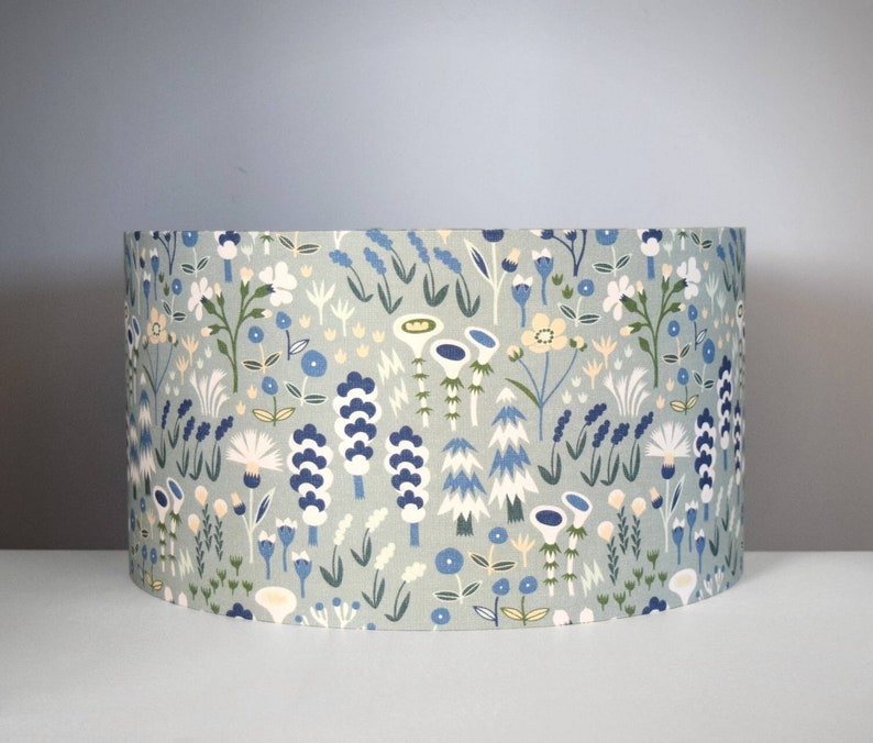 Puede incluir: Una pantalla de l&aacute;mpara cil&iacute;ndrica con un estampado floral. El dise&ntilde;o presenta flores y follaje estilizados en tonos azules, blancos y verdes sobre un fondo verde claro. Un art&iacute;culo de decoraci&oacute;n para el hogar.