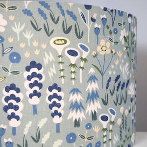Puede incluir: Pantalla de l&aacute;mpara cil&iacute;ndrica con fondo azul claro y un patr&oacute;n repetido de flores y follaje estilizados en tonos de azul, blanco, verde y amarillo. El dise&ntilde;o evoca un tema bot&aacute;nico caprichoso.