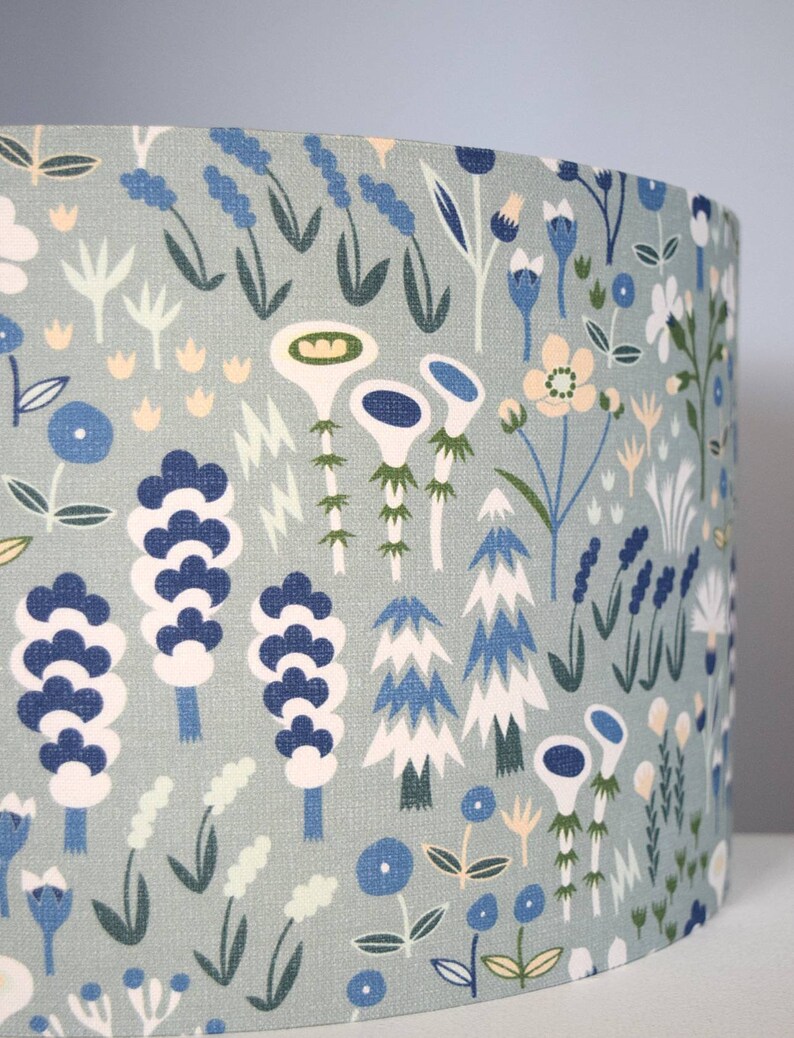 Puede incluir: Pantalla de l&aacute;mpara cil&iacute;ndrica con fondo azul claro y un patr&oacute;n repetido de flores y follaje estilizados en tonos de azul, blanco, verde y amarillo. El dise&ntilde;o evoca un tema bot&aacute;nico caprichoso.