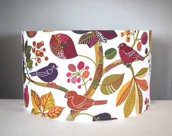 May include: A cylindrical lampshade featuring a white background with a repeating pattern of birds, leaves, and berries. The design includes birds in shades of purple and rust, with foliage in yellow, green, and dark red, creating a dense botanical motif.
