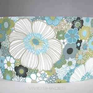 May include: Cylindrical lampshade featuring a floral pattern. The design includes large white flowers with blue centers, complemented by smaller blue, green, and black floral accents. The text "VIVID SHADES" is visible at the bottom.