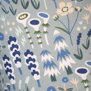 Puede incluir: Primer plano de una tela con un estampado floral repetido. El dise&ntilde;o presenta flores y follaje estilizados en tonos de azul, blanco, verde y melocot&oacute;n sobre un fondo gris azulado claro. El patr&oacute;n tiene un estilo caprichoso e ilustrativo.