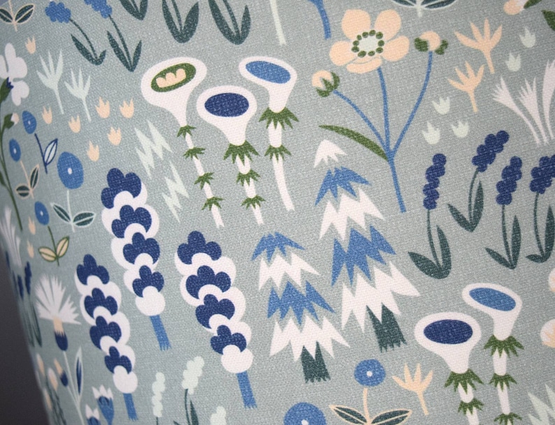 Puede incluir: Primer plano de una tela con un estampado floral repetido. El dise&ntilde;o presenta flores y follaje estilizados en tonos de azul, blanco, verde y melocot&oacute;n sobre un fondo gris azulado claro. El patr&oacute;n tiene un estilo caprichoso e ilustrativo.