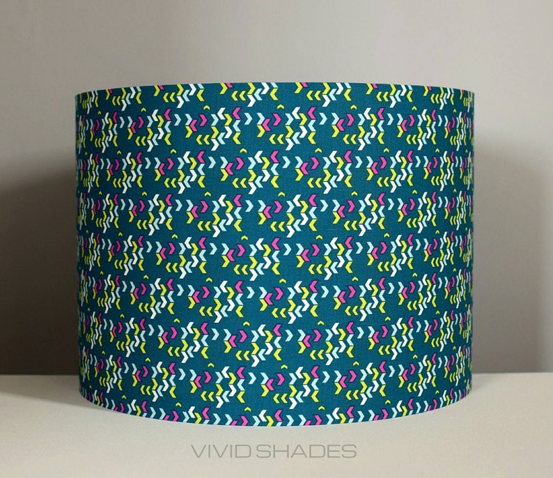 May include: Teal lampshade with a geometric pattern of white, yellow, and pink arrows. The lampshade has the text "VIVID SHADES" printed on the bottom.