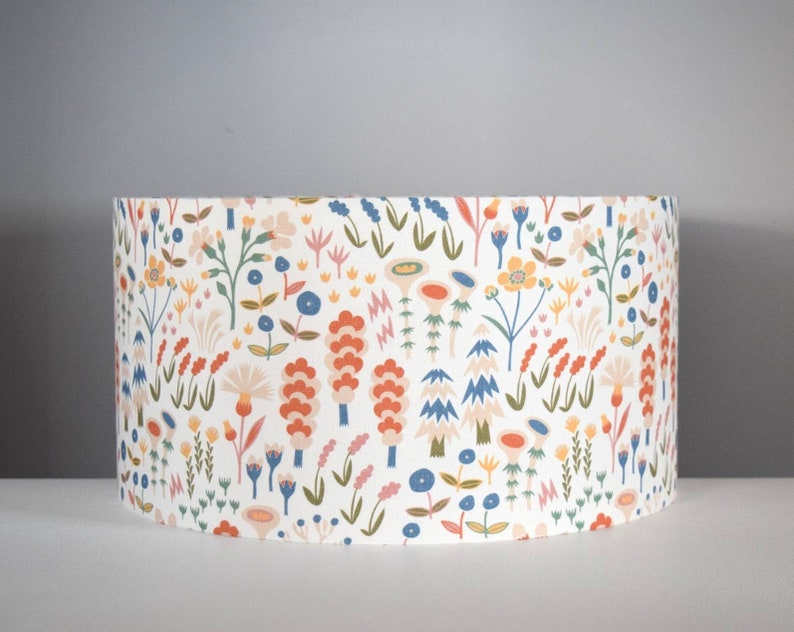 Puede incluir: Pantalla de l&aacute;mpara cil&iacute;ndrica con un estampado floral colorido sobre fondo blanco. El dise&ntilde;o incluye diversas flores en tonos naranja, azul, amarillo y rosa, con tallos y hojas verdes. Un elemento decorativo para el hogar.