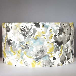 May include: A cylindrical lampshade with a watercolour floral design. The pattern features splashes of yellow, grey, and blue on a white background. The lampshade is a home decor item.