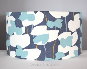 May include: A cylindrical lampshade with a floral pattern. The design features large white and light blue flower shapes against a dark blue background. Thin yellow and light blue lines add detail. The text "VIVID SHADES" is at the bottom.