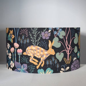 Puede incluir: Pantalla de l&aacute;mpara cil&iacute;ndrica con un patr&oacute;n repetido de una liebre corriendo y elementos florales. El dise&ntilde;o presenta una liebre con un patr&oacute;n a cuadros amarillo y naranja, sobre un fondo azul oscuro con flores y follaje coloridos.