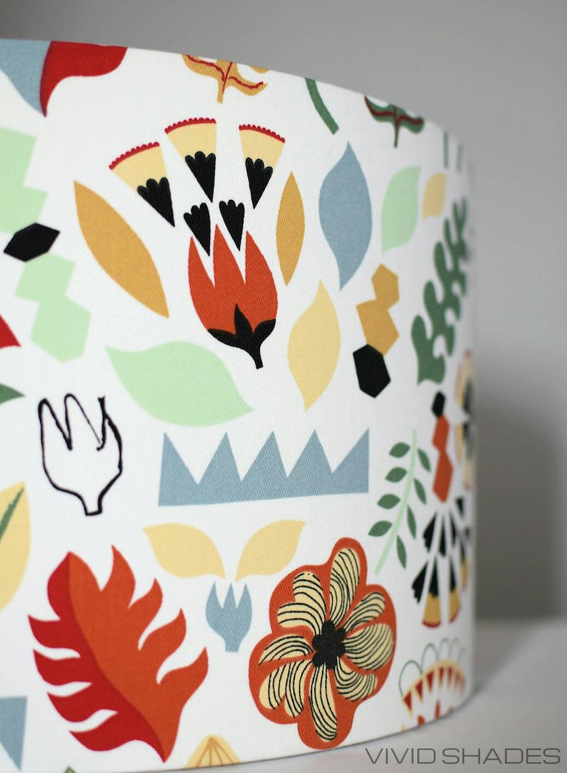 May include: A white lampshade with a colorful floral pattern. The pattern features red, orange, yellow, green, and blue flowers and leaves. The lampshade has the text "VIVID SHADES" printed on the bottom.