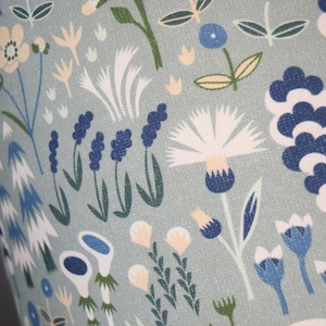Puede incluir: Primer plano de una tela con un estampado floral repetido. El dise&ntilde;o presenta flores y follaje estilizados en tonos de azul, blanco y crema sobre un fondo verde apagado. El patr&oacute;n tiene un estilo caprichoso e ilustrativo.