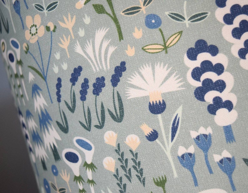 Puede incluir: Primer plano de una tela con un estampado floral repetido. El dise&ntilde;o presenta flores y follaje estilizados en tonos de azul, blanco y crema sobre un fondo verde apagado. El patr&oacute;n tiene un estilo caprichoso e ilustrativo.