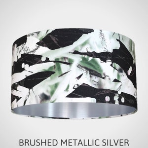 May include: A lampshade with a black, white, and green abstract pattern. The bottom of the lampshade is a brushed metallic silver.