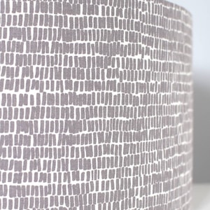 May include: A cylindrical lampshade featuring a repeating pattern of grey rectangular shapes on a white backdrop. The fabric has a textured appearance, and the design is evenly spaced across the surface.