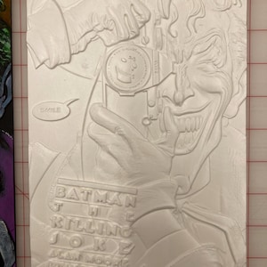May include: A white relief sculpture of the Joker from the Batman comic "The Killing Joke." The Joker is holding a camera and smiling widely. The word "SMILE" is in a speech bubble. The comic title and creators are listed.