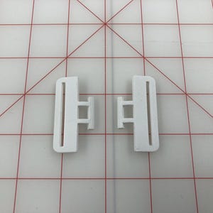 以下が含まれることがあります： 長方形のフレームと中央のバーを備えた、2つの白いプラスチック製のサイドリリースバックル。バックルはストラップを接続して固定するように設計されています。背景はグリッドパターンです。