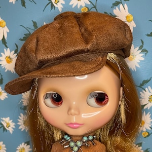 May include: A doll with long blonde hair, wearing a brown newsboy cap, a brown sweater, and a color-block skirt with orange and brown panels. The doll has a turquoise necklace and is set against a blue floral background.