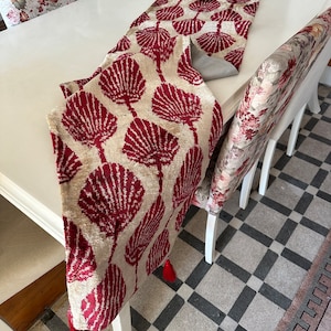 May include: A red and cream table runner with a leaf pattern and tassels on the ends. The runner is on a white table with floral patterned chairs.