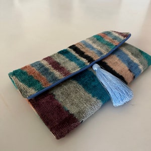 May include: A colorful striped clutch with a blue trim and a light blue tassel. The clutch is made of a soft, velvety material.