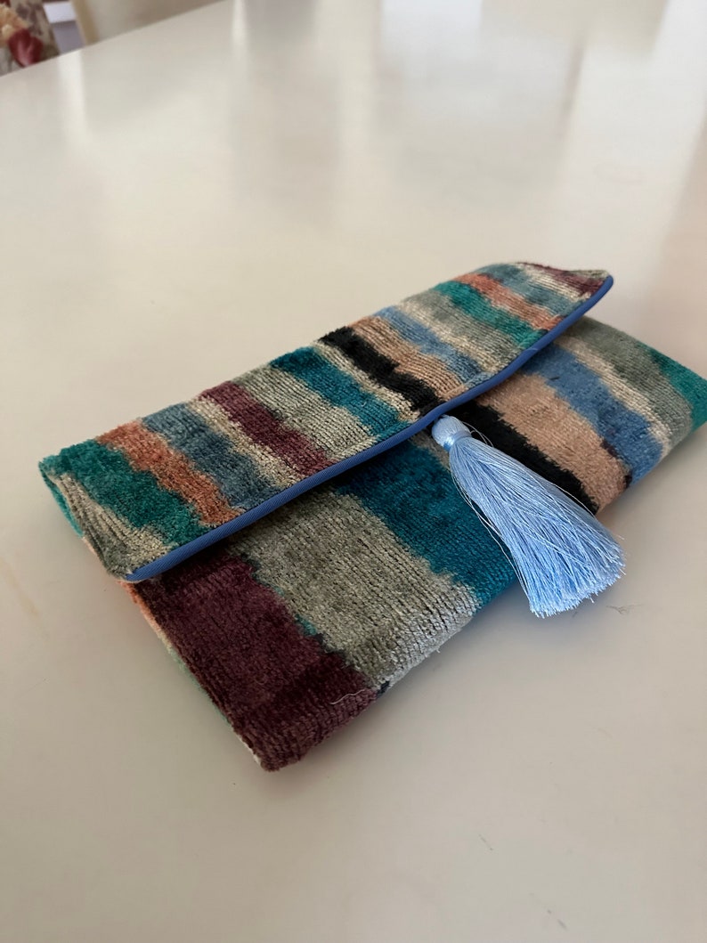 May include: A colorful striped clutch with a blue trim and a light blue tassel. The clutch is made of a soft, velvety material.