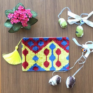 Op de afbeelding: Een kleurrijke clutch met een geometrisch patroon en een gele kwast. De tas ligt op een houten oppervlak met verschillende kleine, met stof beklede voorwerpen en een wit lint.