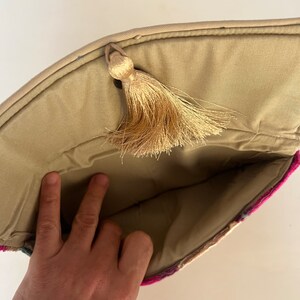 Może przedstawiać: Beżowa torebka typu clutch z zapięciem na frędzle. Torebka jest podszyta beżową tkaniną i ma kolorowy wz&oacute;r na zewnątrz.