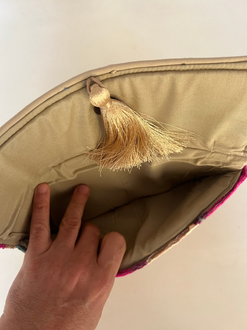 Może przedstawiać: Beżowa torebka typu clutch z zapięciem na frędzle. Torebka jest podszyta beżową tkaniną i ma kolorowy wz&oacute;r na zewnątrz.