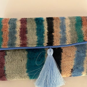 May include: A striped clutch purse with a blue tassel. The purse is made of a soft, textured fabric and has a blue trim.