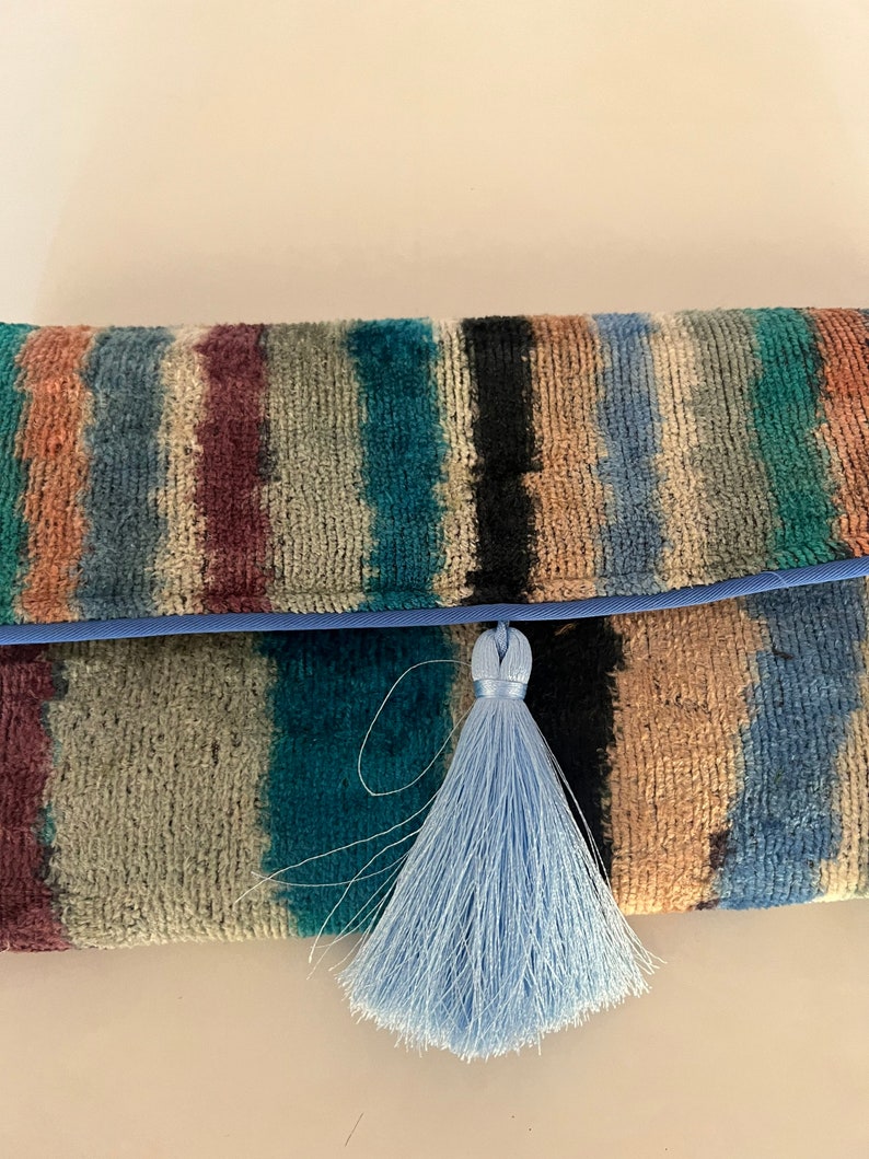 May include: A striped clutch purse with a blue tassel. The purse is made of a soft, textured fabric and has a blue trim.